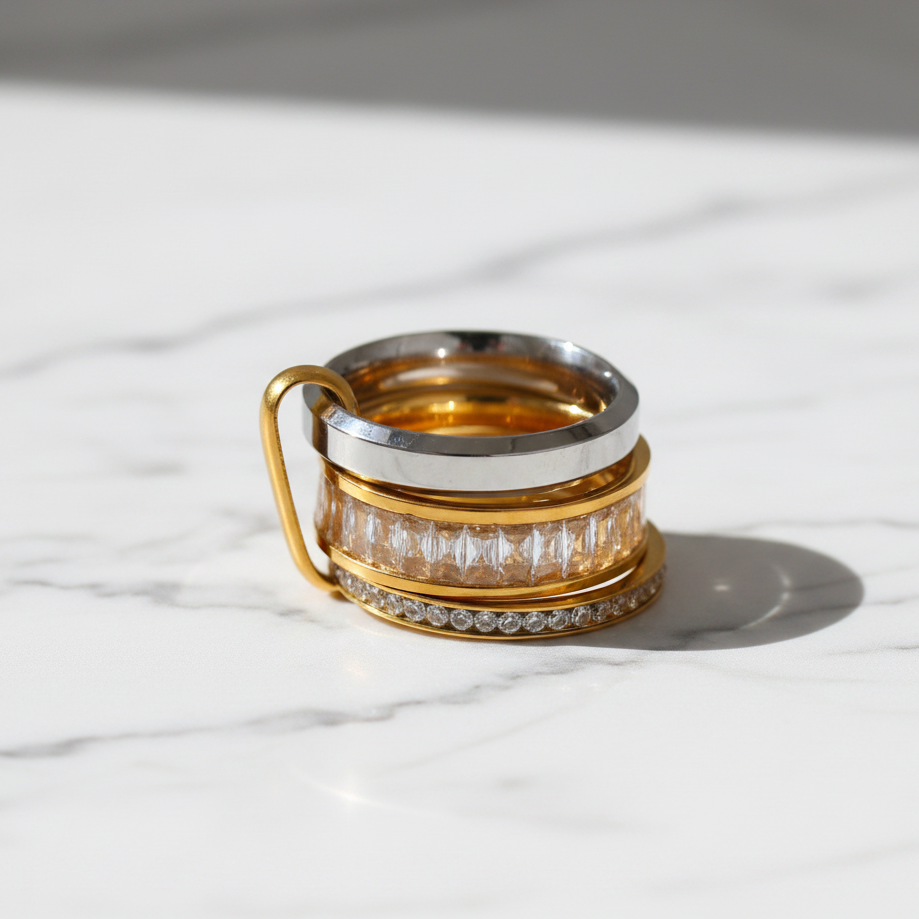 A stack of three rings on a white background, featuring gold and silver metal bands with clear gemstones. The top ring is a polished silver band, the middle ring is a gold band with a row of clear rectangular-cut stones, and the bottom ring is a gold band with a row of clear round-cut stones.
