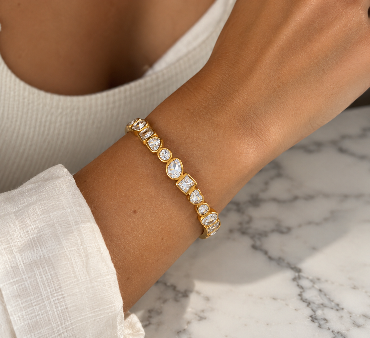 A gold bracelet with multiple clear round and baguette-cut gemstones worn on a person's wrist, shown against a white top and a white marble surface.