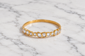 A gold band ring featuring a row of clear marquise and heart-shaped gemstones, displayed on a white and gray marble surface.