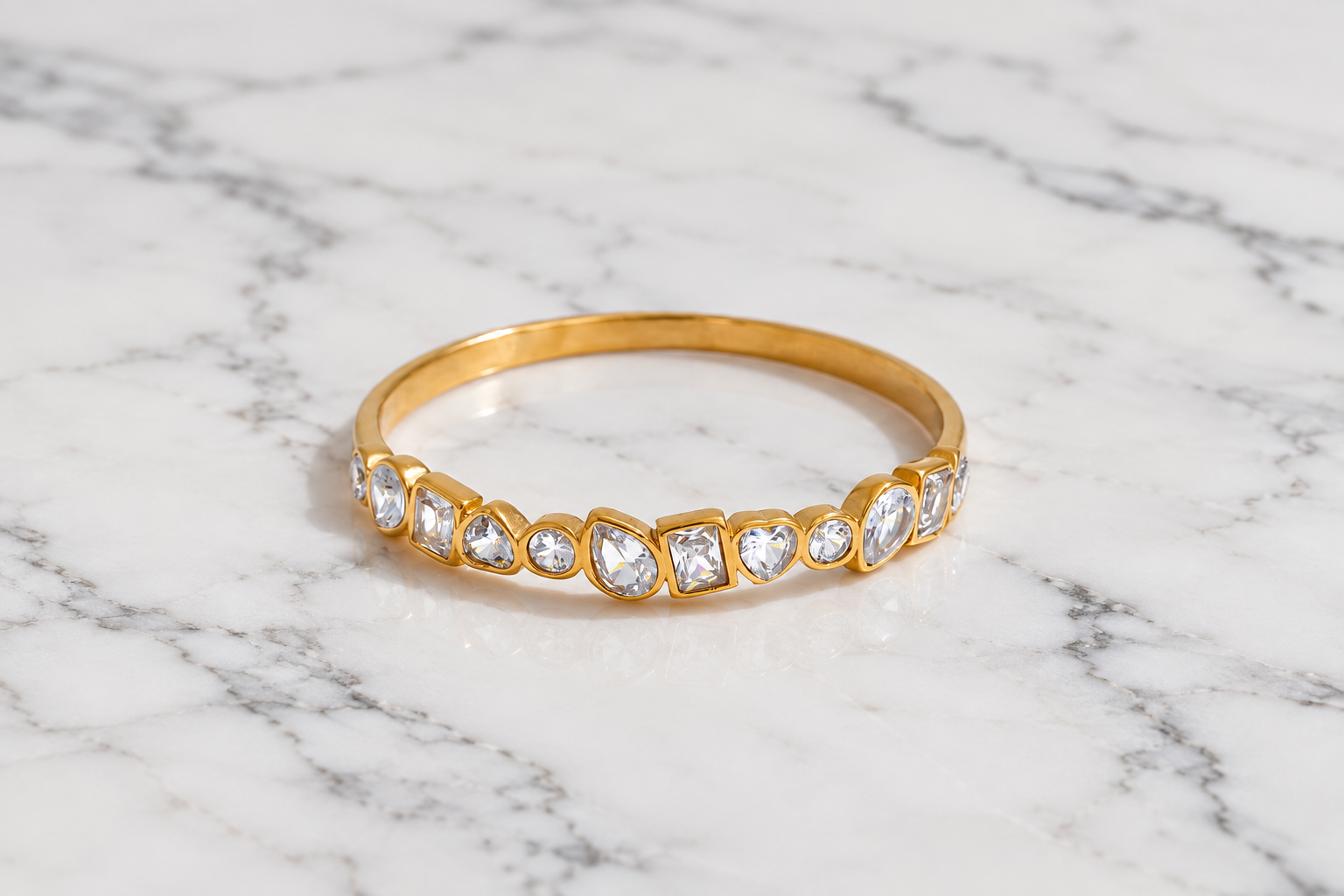 A gold band ring featuring a row of clear marquise and heart-shaped gemstones, displayed on a white and gray marble surface.