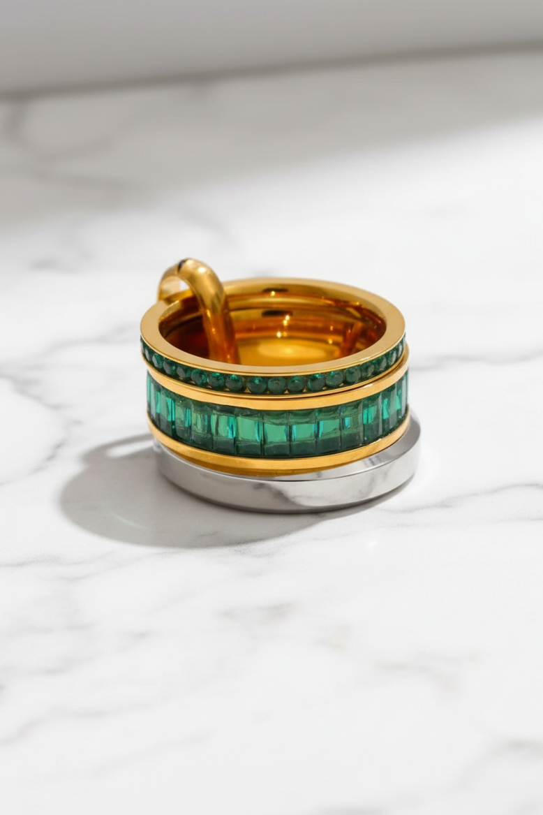 A luxury gold ring with a double row of emerald-cut green stones, displayed on a white marble surface with a soft shadow.