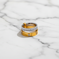 A stack of three rings displayed on a white background within a larger dark interface frame. The rings include a yellow gold band with small clear stones, a silver band with a row of clear stones, and a gold band with a clasp and small round stones.