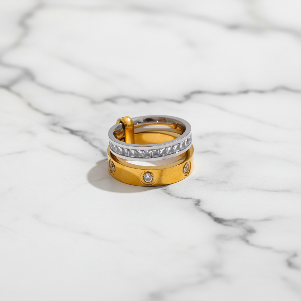 A stack of three rings displayed on a white background within a larger dark interface frame. The rings include a yellow gold band with small clear stones, a silver band with a row of clear stones, and a gold band with a clasp and small round stones.