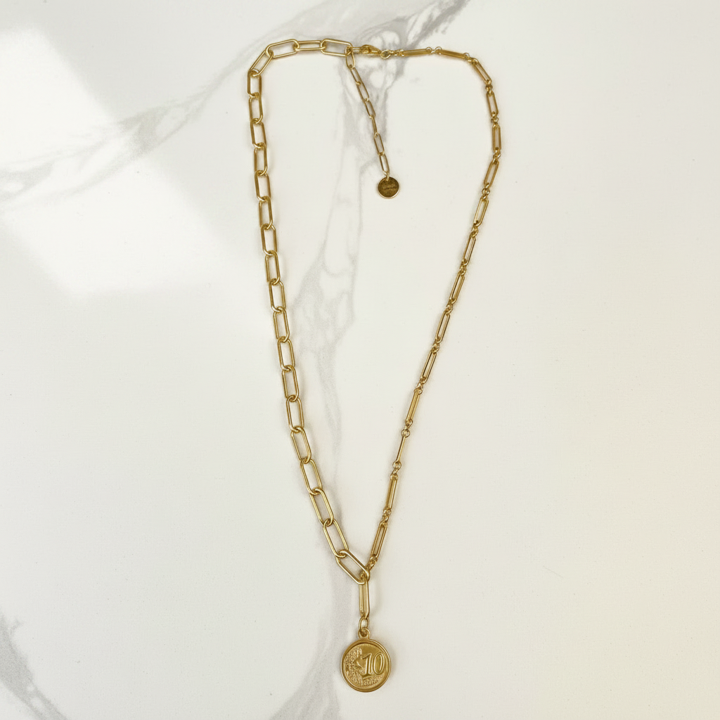 A gold chain necklace with a double-layered link design and a round coin pendant, displayed on a light marble background.