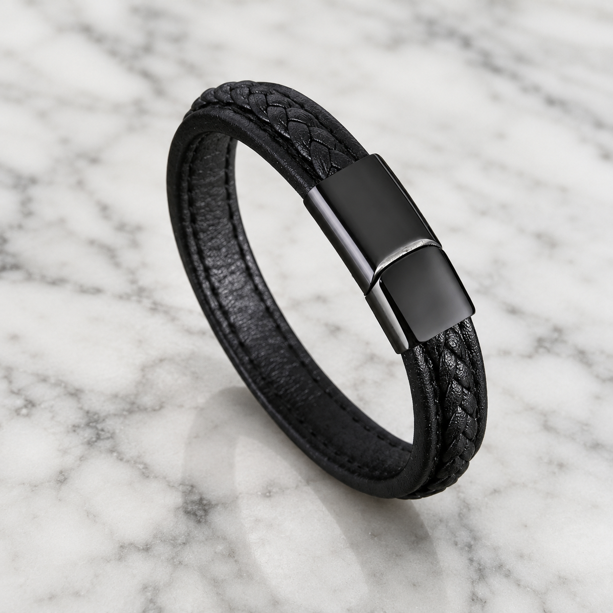 A black braided leather bracelet with a matte black metal clasp, displayed on a white and gray marble surface.